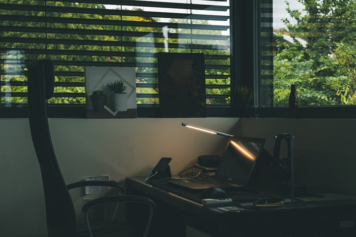 A laptop glowing on a dark desk in a dim room, daylight coming through half-open venetian blinds