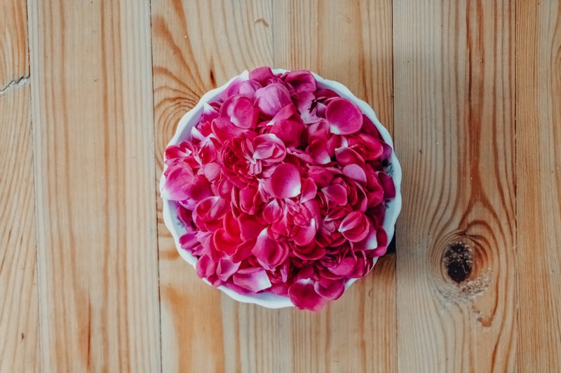 A white ceramic bowl filled with pink rose petals on a pale wooden table