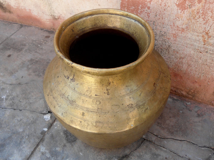A weathered brass water pot resting on a stone floor against a sun-worn terracotta wall