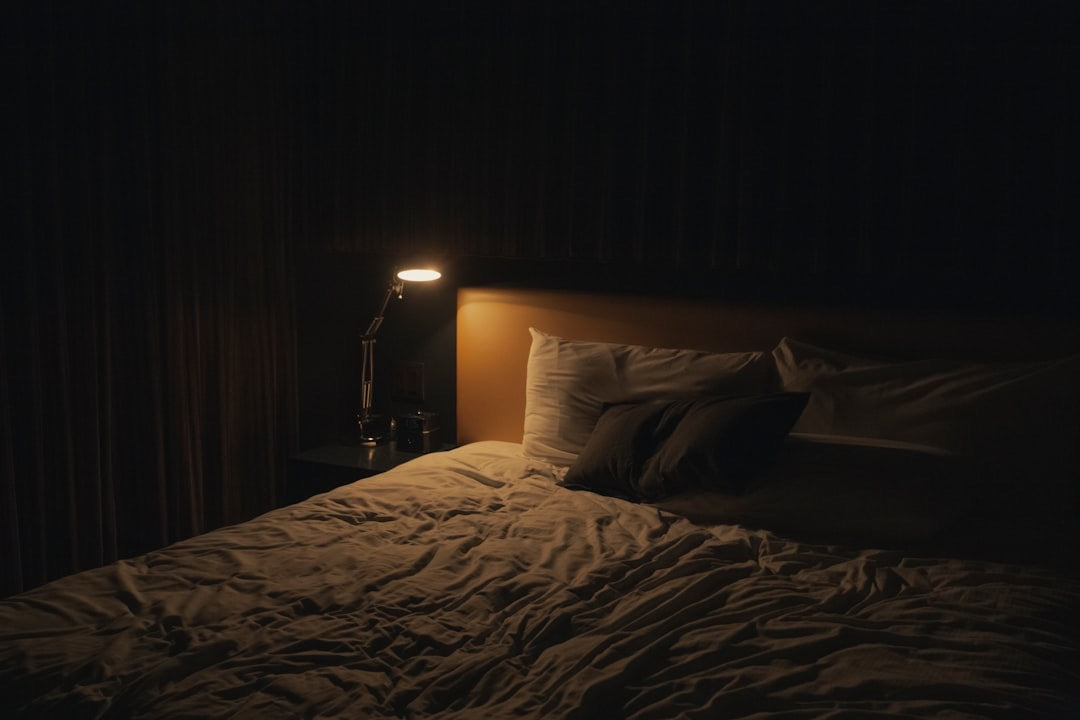 An empty bed with rumpled linen in a dark room, lit only by an adjustable reading lamp on the bedside table