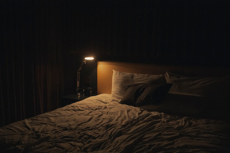 An empty bed with rumpled linen in a dark room, lit only by an adjustable reading lamp on the bedside table