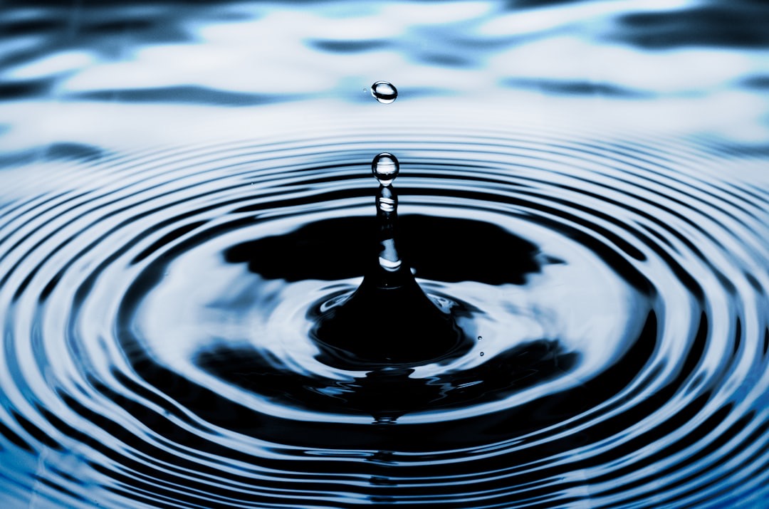 A single water droplet rebounding from the surface of still water, concentric ripples spreading out around it.