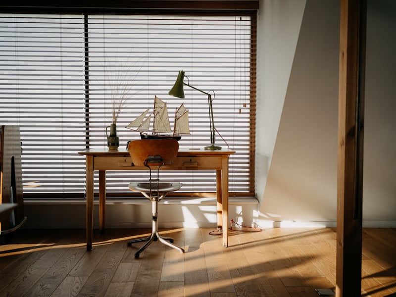 A wooden desk and empty chair beneath a window with half-open blinds in afternoon light