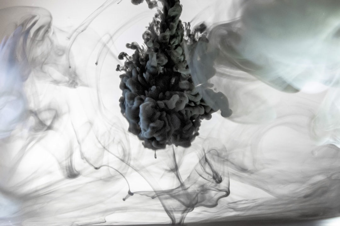 Dark ink billowing through clear water, a dense cloud at the centre with fine tendrils trailing away through the pale liquid.